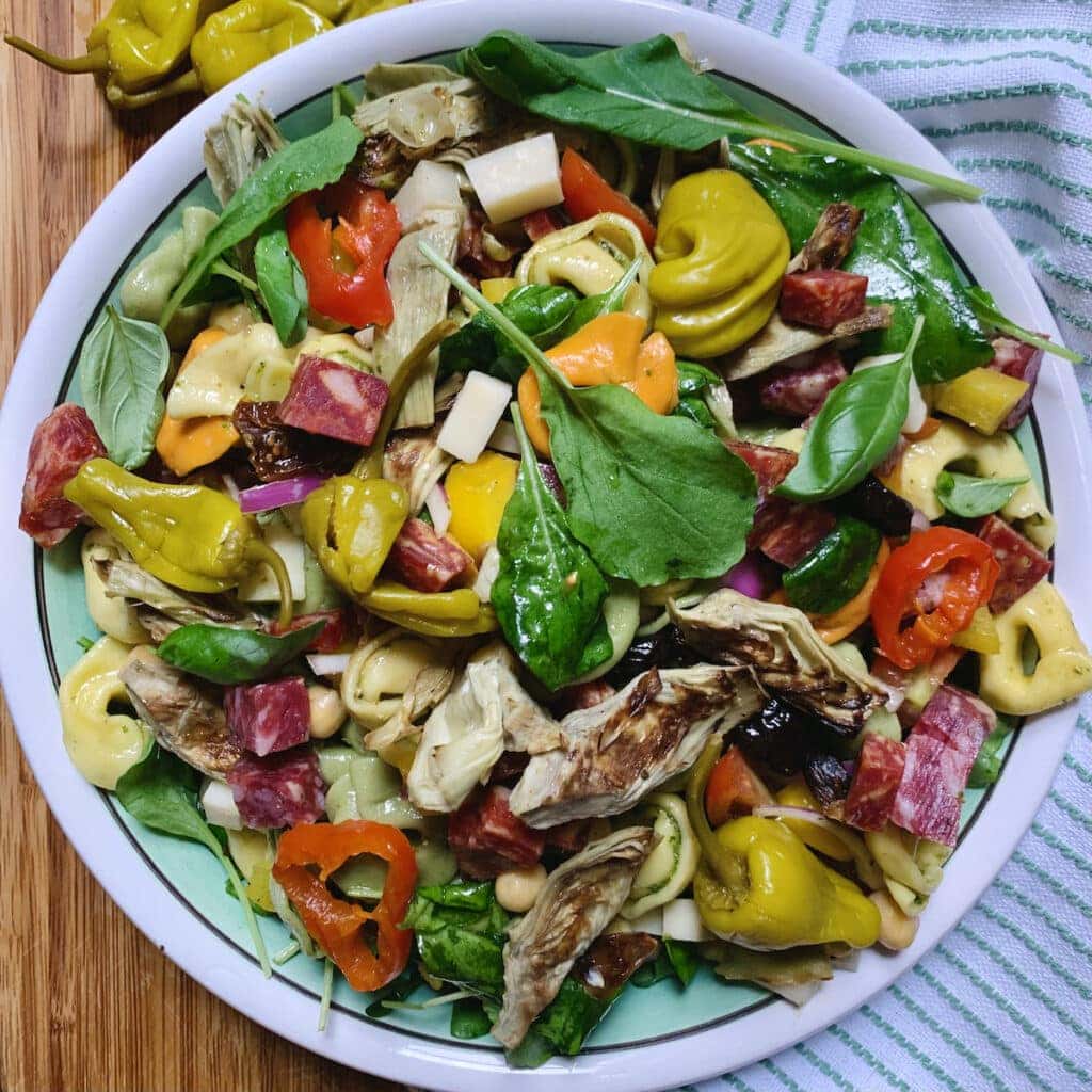 Antipasto tortellini pasta salad in decorative bowl on a white and green towel with pickled peppers in background.