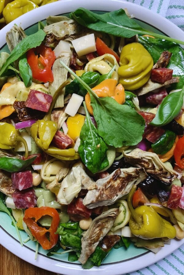 Antipasto tortellini pasta salad in decorative bowl on a white and green towel with pickled peppers in background.