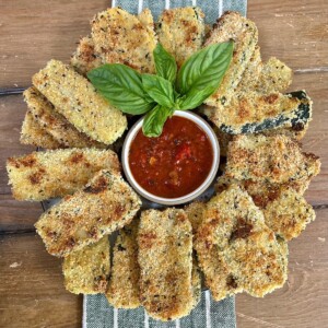 Beautiful arrangement of baked panko crusted zucchini on a round platter with a small bowl of tomato sauce in the center with basil garnish.