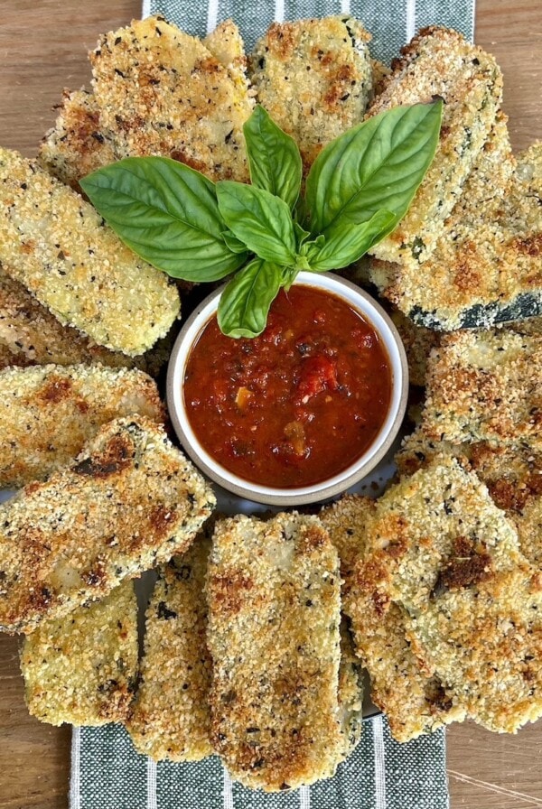 Beautiful arrangement of baked panko crusted zucchini on a round platter with a small bowl of tomato sauce in the center with basil garnish.