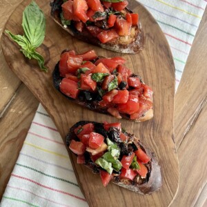 Three pieces of authentic bruschetta di pomodoro with olive on cutting board on towel with basil garnish.