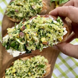 Close up in hand of one piece of zucchini pesto bruschetta with more pieces in background.