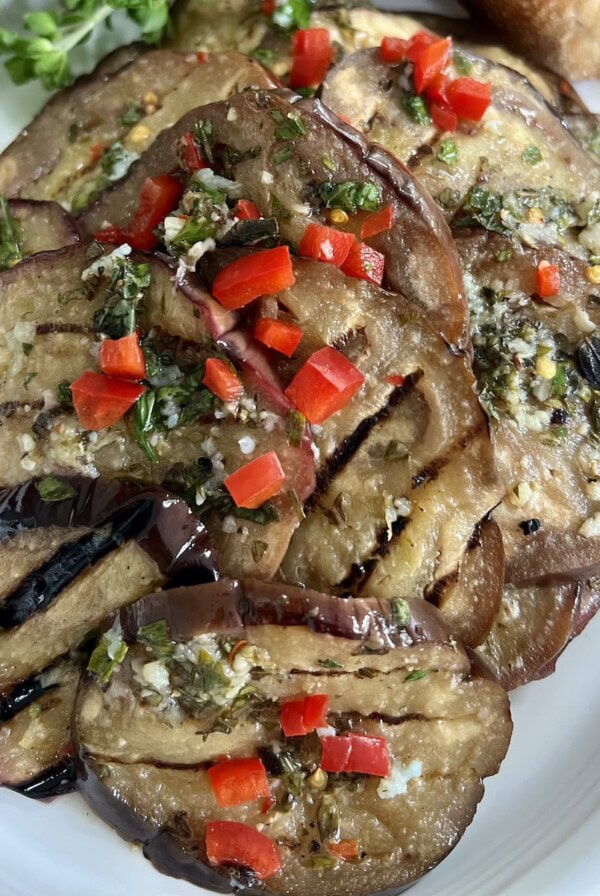 Italian grilled eggplant on white oval plate with toasted bread and fresh herbs in background.
