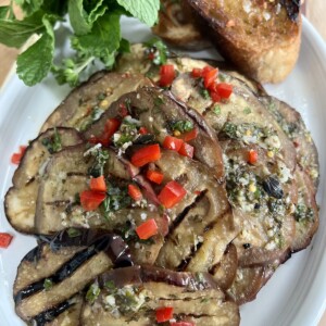 Italian grilled eggplant on white oval plate with toasted bread and fresh herbs in background.