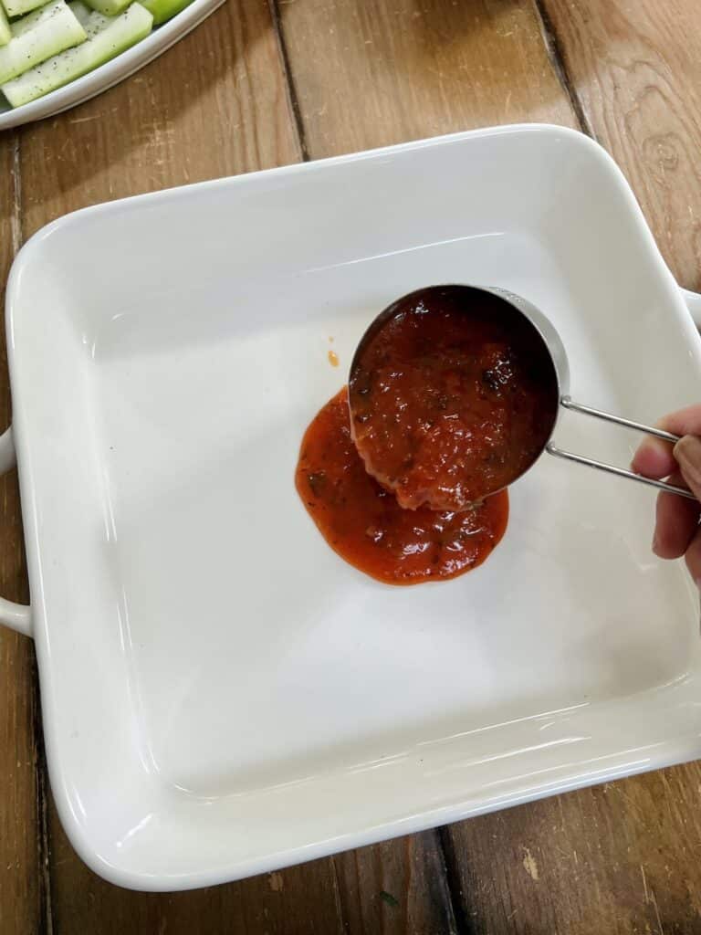 Pouring some tomato sauce into white baking dish with a measuring cup.