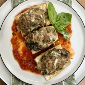 Three pieces of Baked Stuffed Cucuzza Squash over tomato sauce on white plate with basil sprig garnish.