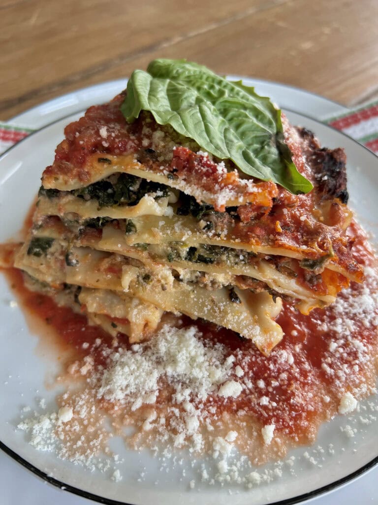 A piece of Lasagna al Forno topped with a basil leaf on a white plate with extra sauce and grated cheese.