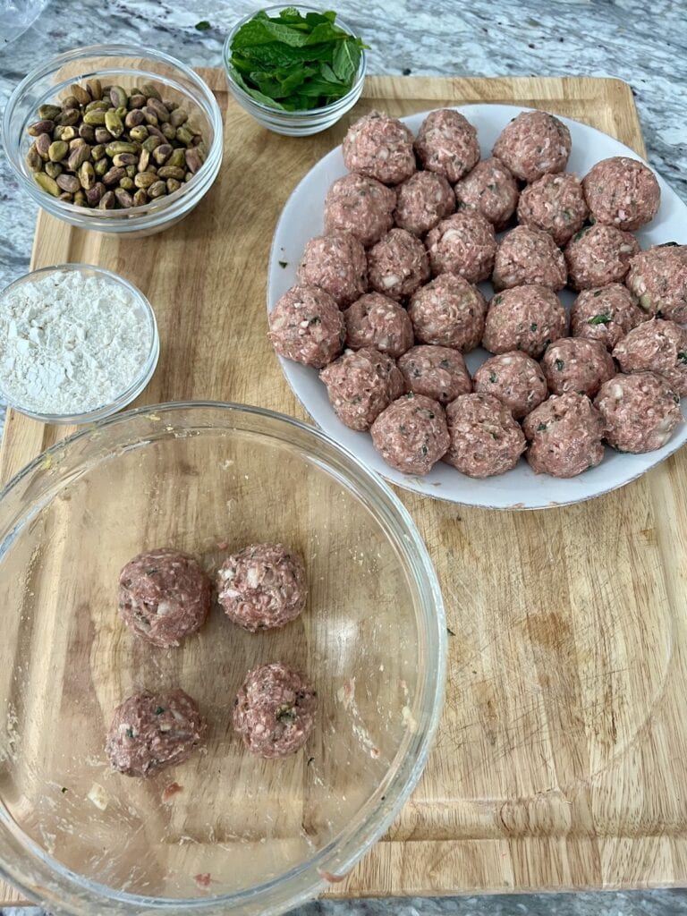 Formed lamb meatballs on white plate with bowls of mint, pistachio and flour in the background.