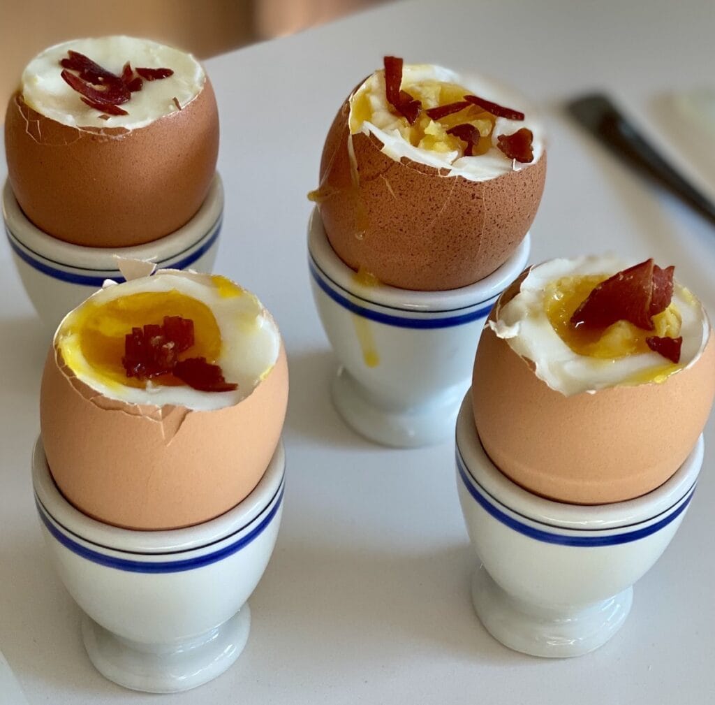 Four soft-boiled eggs in egg holders topped off with some crumbled crispy prosciutto.
