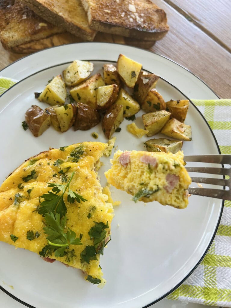 Close up of piece of cheese and ham frittata on fork with the rest of frittata slice and roasted potatoes in background.