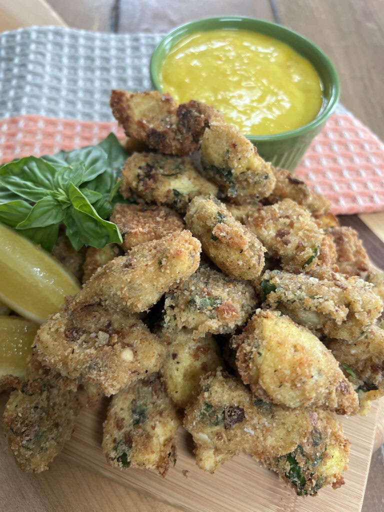 Pan fried artichoke hearts stacked on a cutting board with lemon wedges, fresh basil sprigs and saffron garlic mayonnaise dipping sauce in the background.
