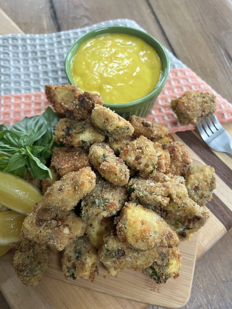 Italian pan fried artichoke hearts stacked on a cutting board with lemon wedges, fresh basil sprigs, saffron garlic mayonnaise dipping sauce and fork in the background.