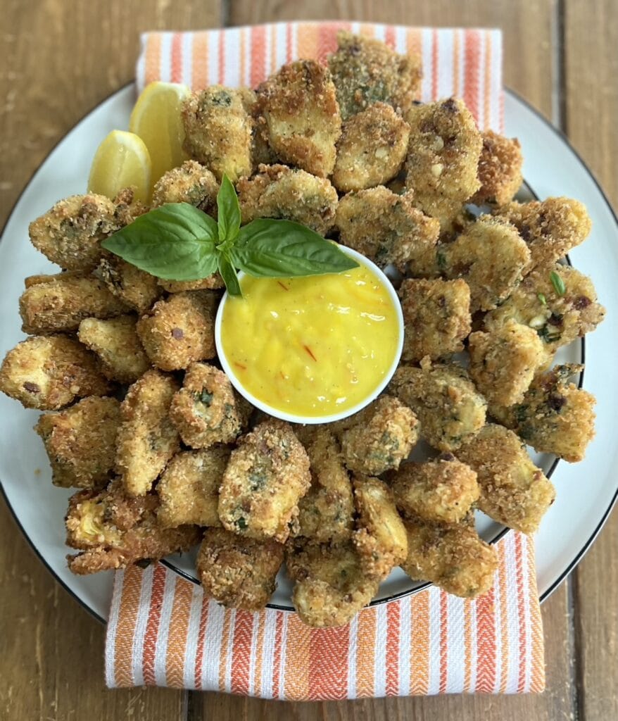 Beautiful white plate full of italian fried artichoke hearts with a bowl of colorful saffron aioli in the center, with basil sprig garnish, over a decorative orange towel.