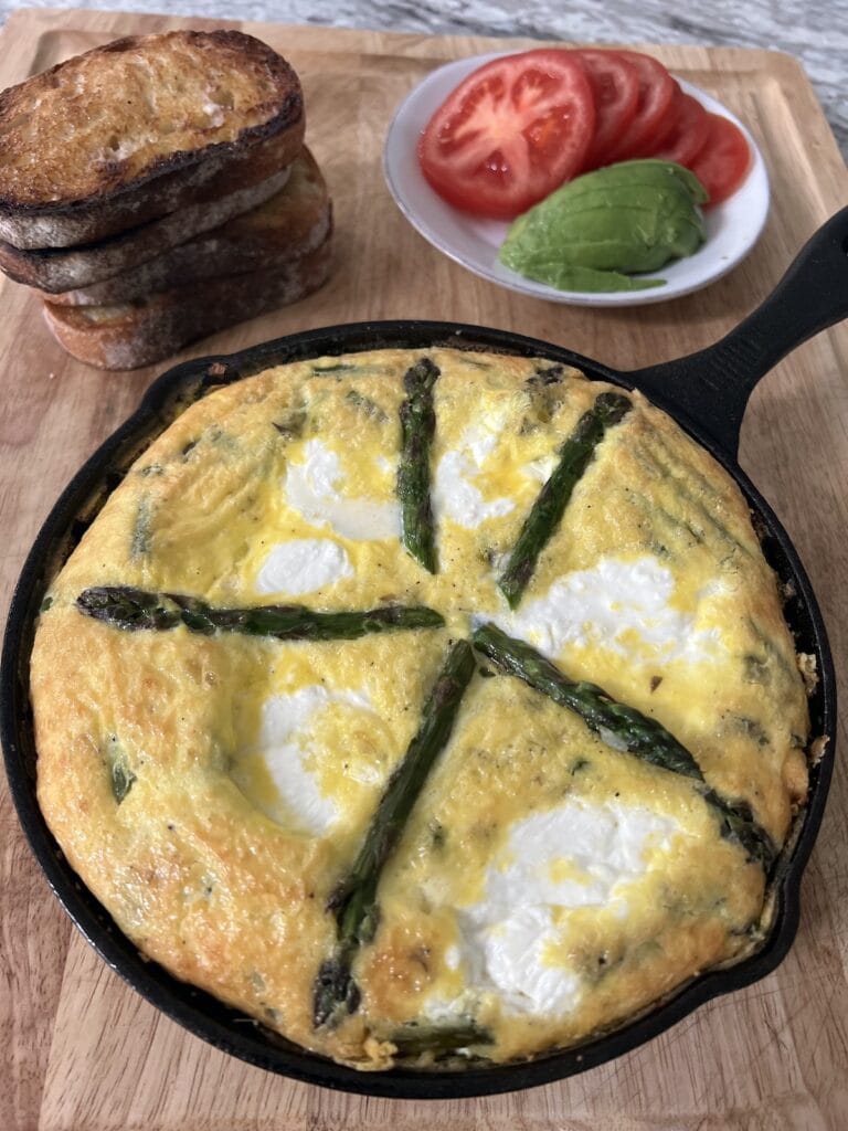 Beautiful asparagus and egg frittata still in cast iron pan on cutting board with a stack of toast and a plate of tomatoes and sliced avocado.