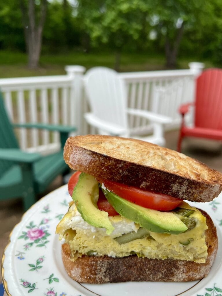 An asparagus and egg frittata sandwich with tomato and avocado on toasted Italian bread, with deck chairs in the background.
