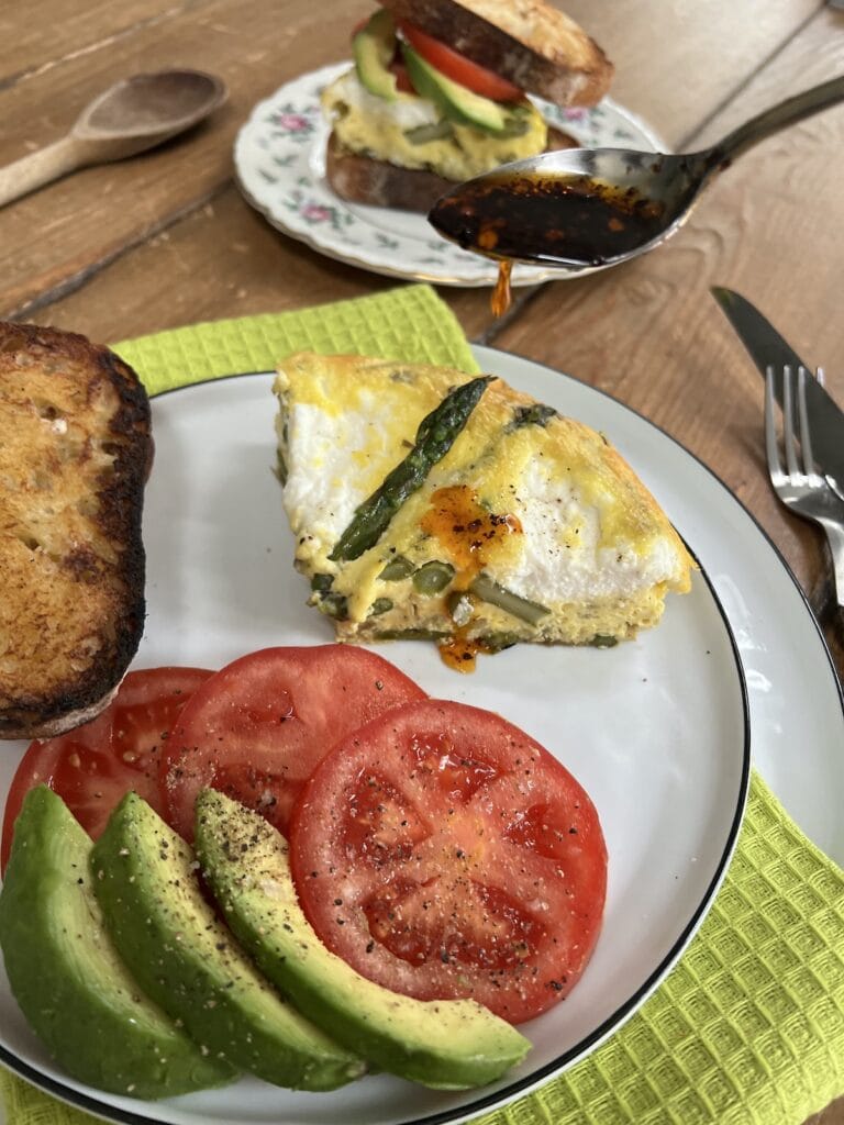Drizzling spicy homemade pepper oil over Italian asparagus and eggs on plate with toast, tomato and avocado, with sandwich in teh background.