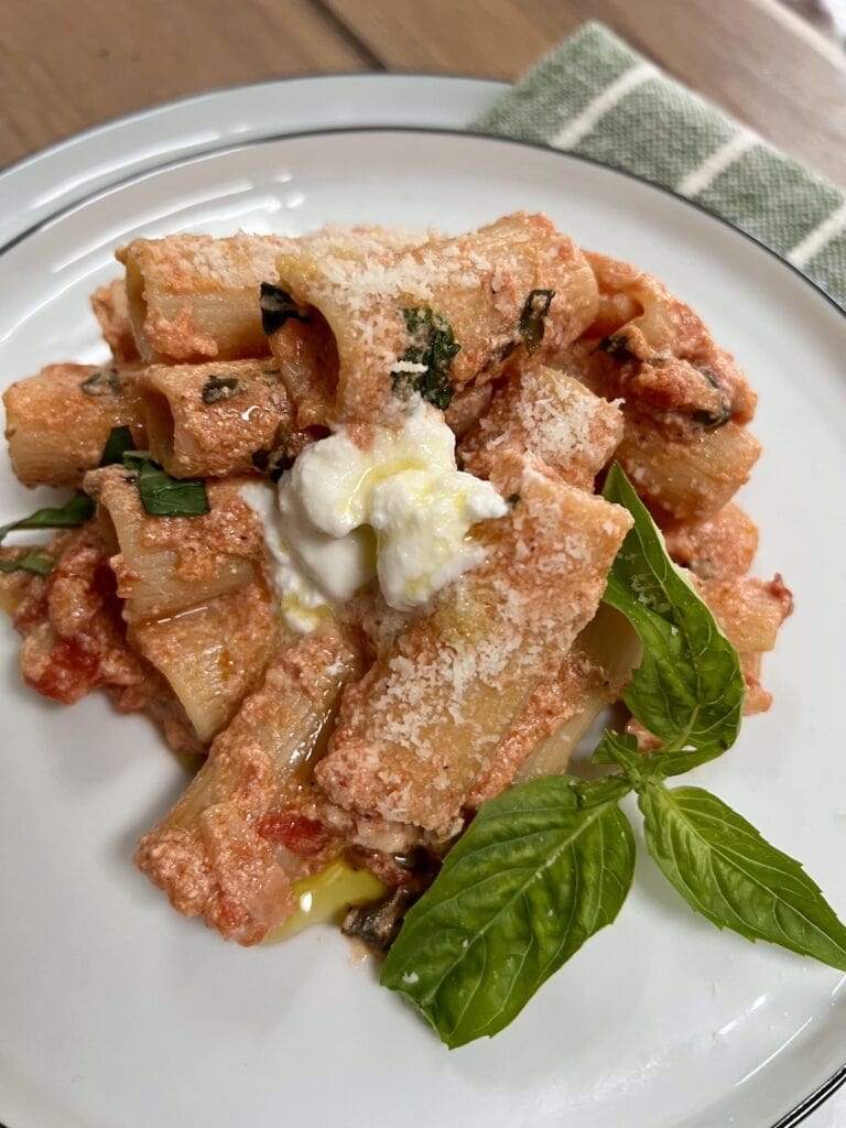 Portion of tomato and ricotta pasta with extra ricotta on white dinner plate with a sprig of basil.
