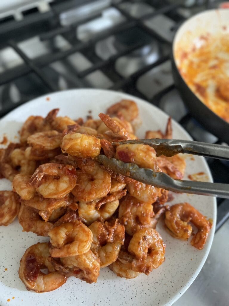 Transferring spicy tomato shrimp from pan to white plate with tongs