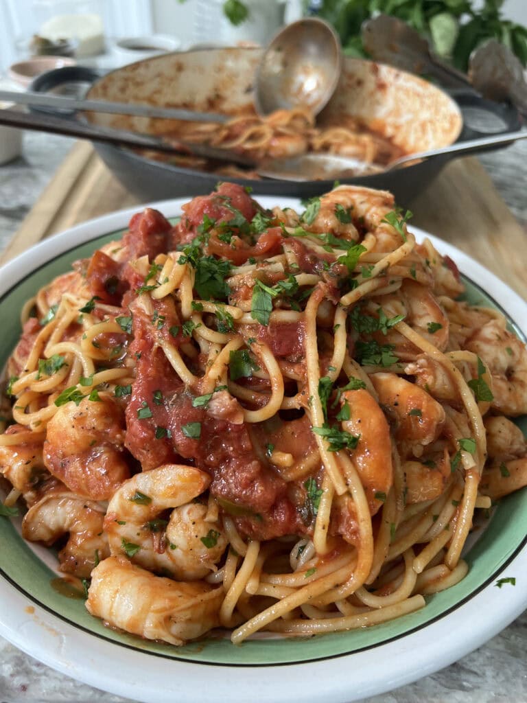 Pasta bowl filled with spicy shrimp marinara with parsley garnish and pan in background with spoon.