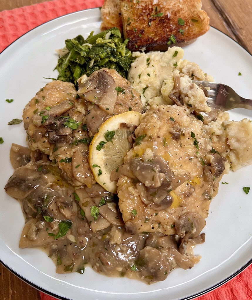 Chicken Francaise with chopped parsley and lemon slice garnish on white dinner plate with mashed potatoes, broccoli rabe, bread and fork.