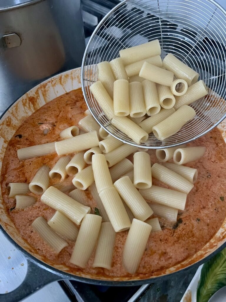 Cooked pasta being added to creamy tomato ricotta sauce in pan.
