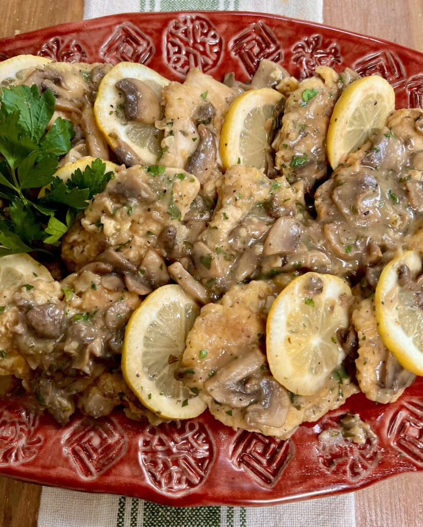 Italian Chicken Francese with Mushrooms with lemon slices and fresh parsley garnish on red serving platter.