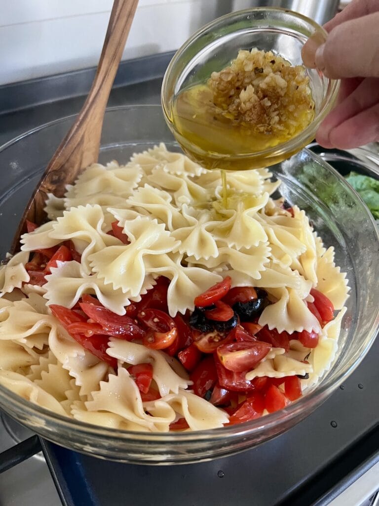 Adding sautéed garlic to bowl with tomato olive mixture and cooked bowtie pasta.
