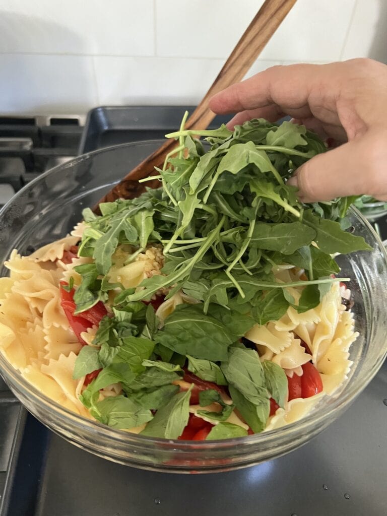 Adding baby arugula to tomato basil pasta salad with bowtie pasta.