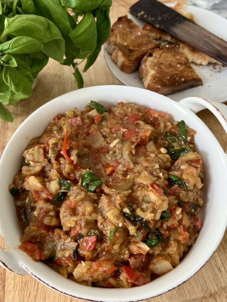 Roasted eggplant and tomato  spread in white serving dish with toast in background.