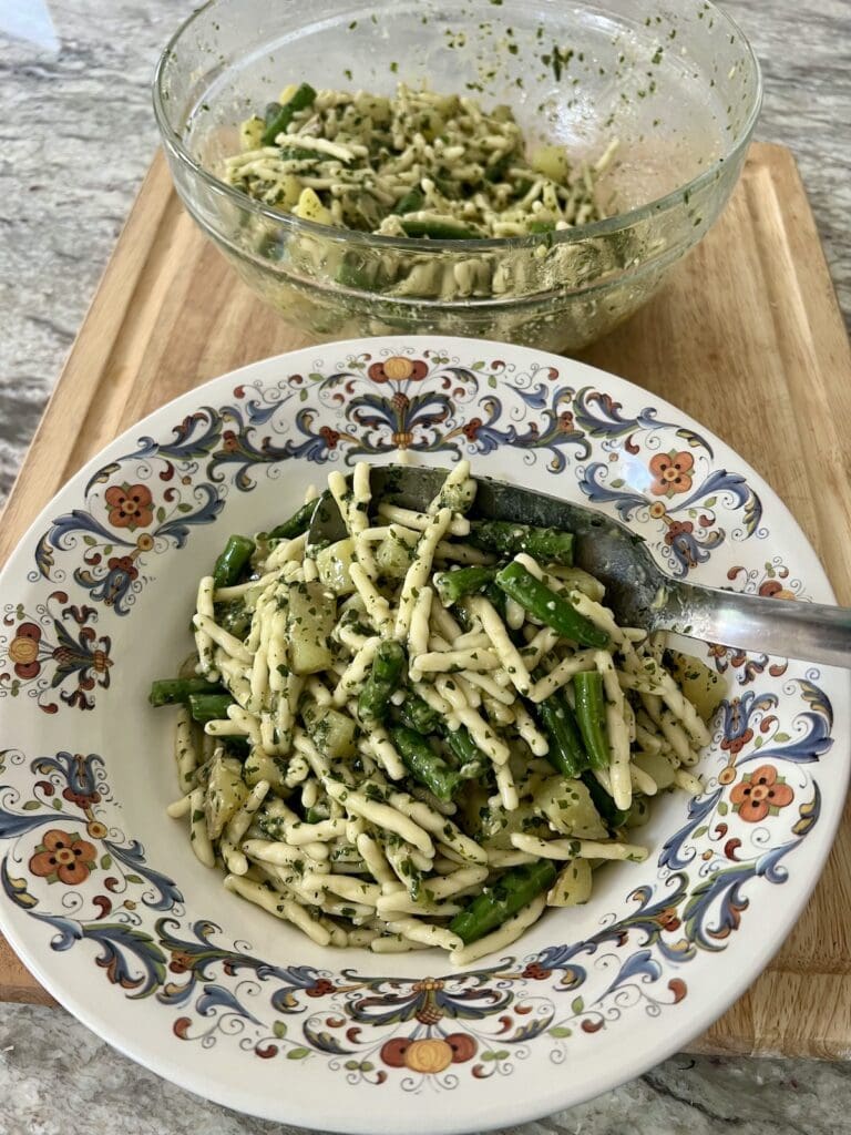 Spooning finished trofie al pesto genovese into serving bowl with large stainless steel spoon.