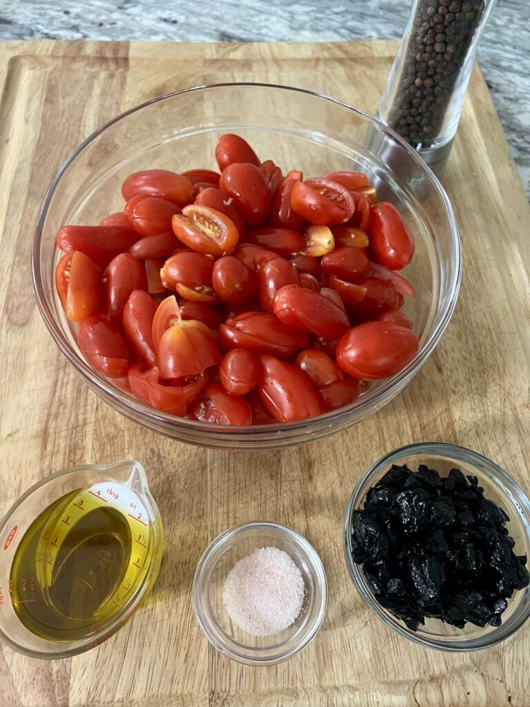 Prepped tomatoes with olive oil, salt and black olives, all in bowls, on cutting board. 