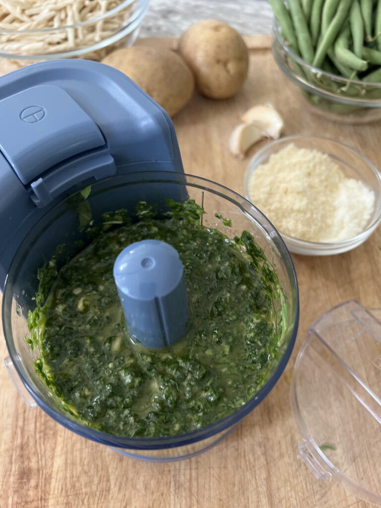 Finished basil pesto in food processor with other ingredients in background.