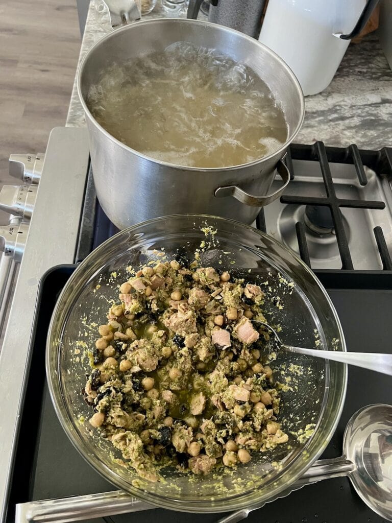 Pasta cooking in large pot of boiling water next to bowl of sauce ingredients.