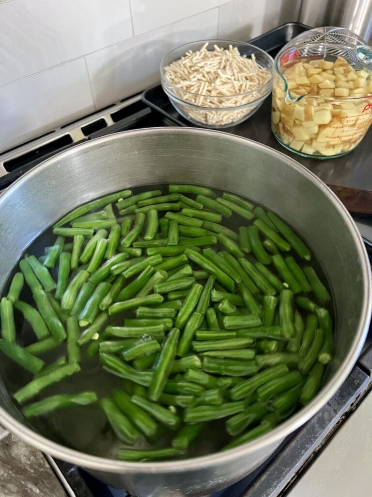 Green beans cooking in large pot with bowls of potatoes and pasta in background.
