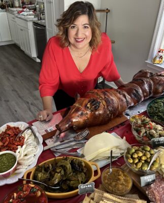 Michele at home in front of table full of food, including a roast pig.
