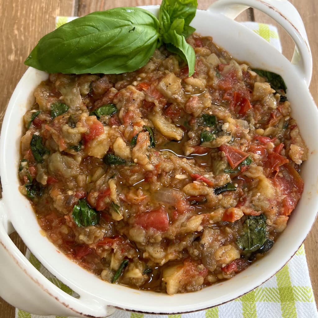 Roasted eggplant and tomato spread in white serving dish with basil garnish.
