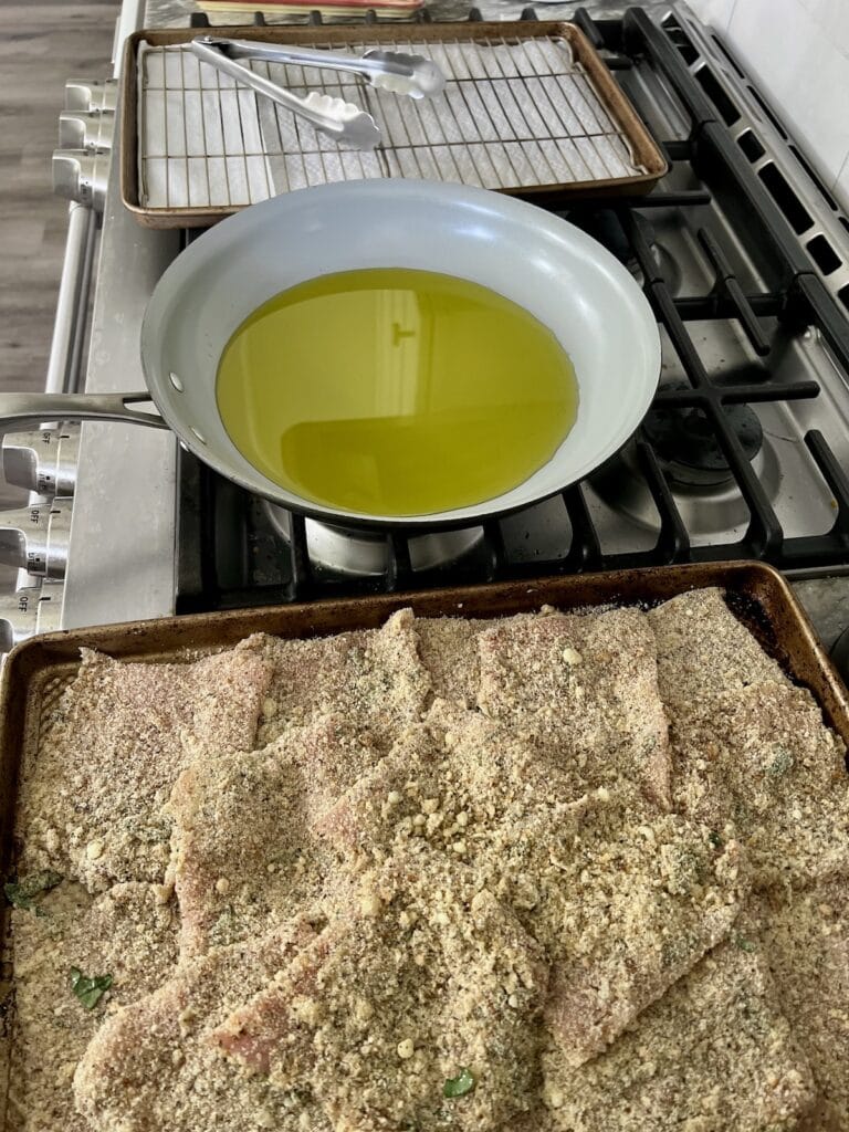 Mis-en-place set-up of pan full of breaded cutlets, next to frying pan with oil next to wire rack over sheet pan, at stove.