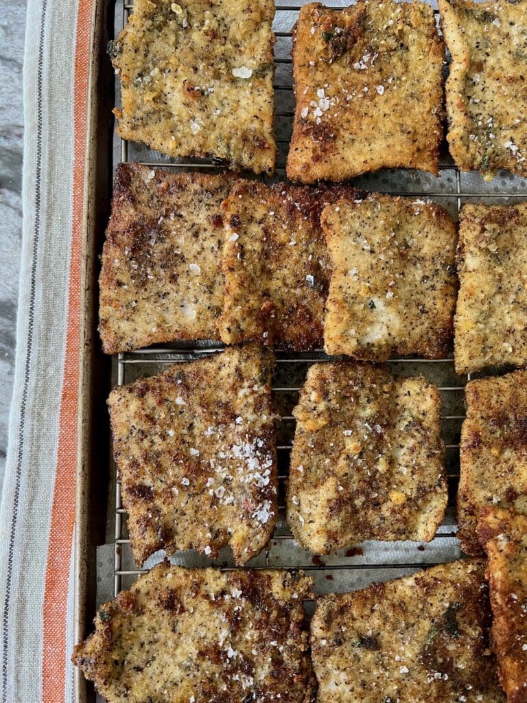 Finished crispy fried Italian pork cutlets on wire rack over sheet pan over a decorative towel.