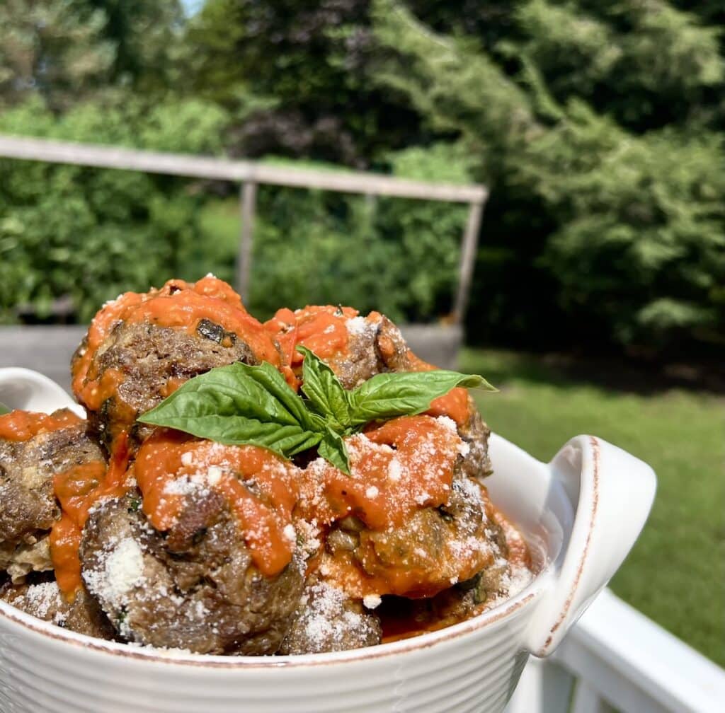 Beef meatballs with eggplant in white serving dish with basil with garden in background.