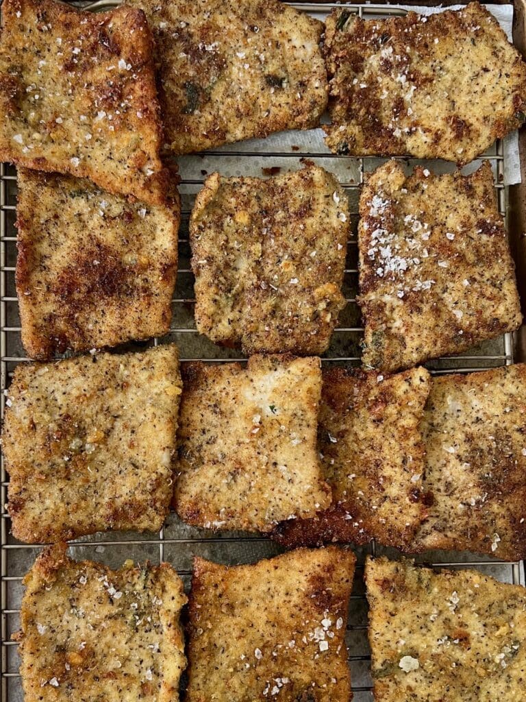 Finished fried pork cutlets sprinled with salt on rack over baking sheet.