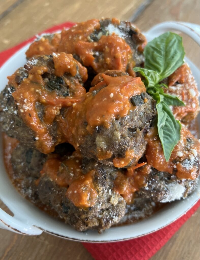 Bowl of eggplant and beef meatballs with basil sprig  drizzled with tomato basil sauce.