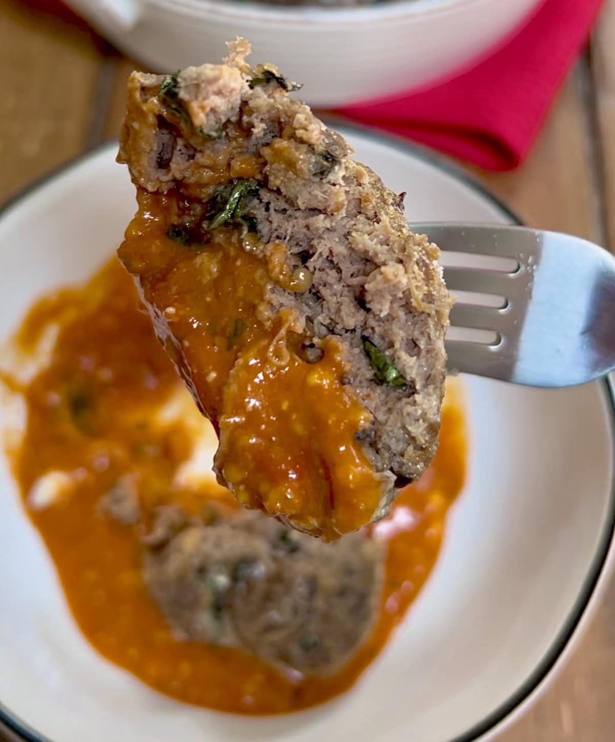 Close up of a meatball half on fork with meatball and sauce in bowl in background.