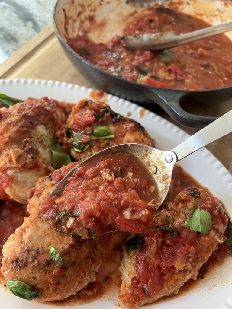 Spooning additional pan sauce over the plated chicken pomodoro in white serving platter.