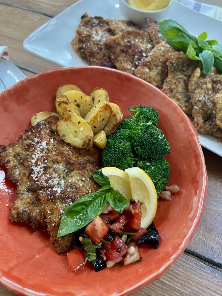 Pork cutlets on orange plate with lemon wedges, basil sprig, steamed broccoli, roasted potatoes and tomato salad with platter of pork milanese in the background.