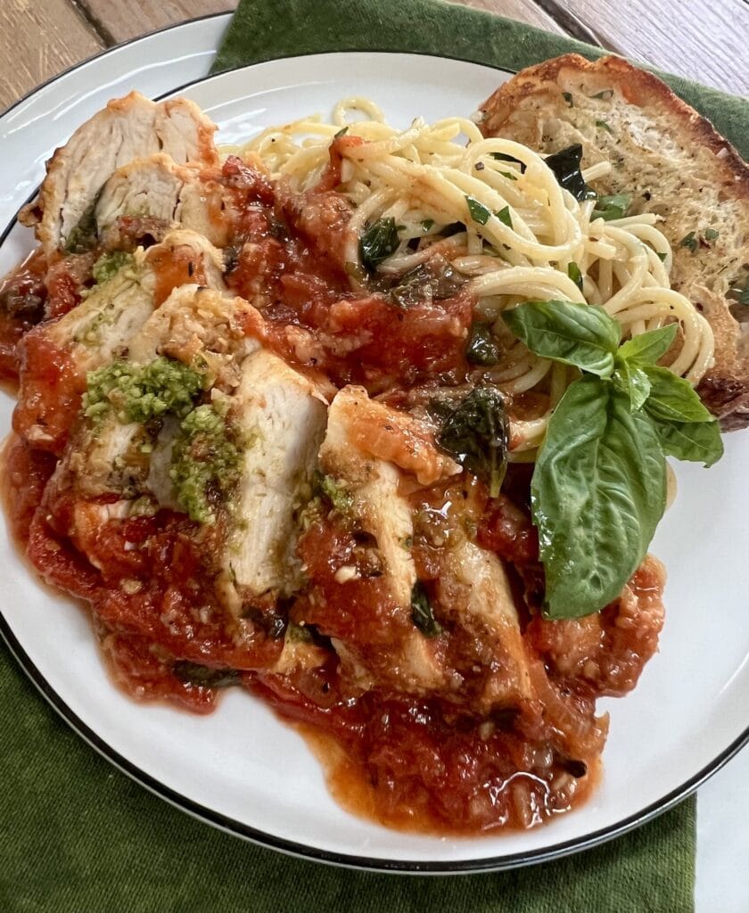 Sliced Pomodoro Chicken on white dinner plate with spaghetti, bread, basil garnish and a little pesto.