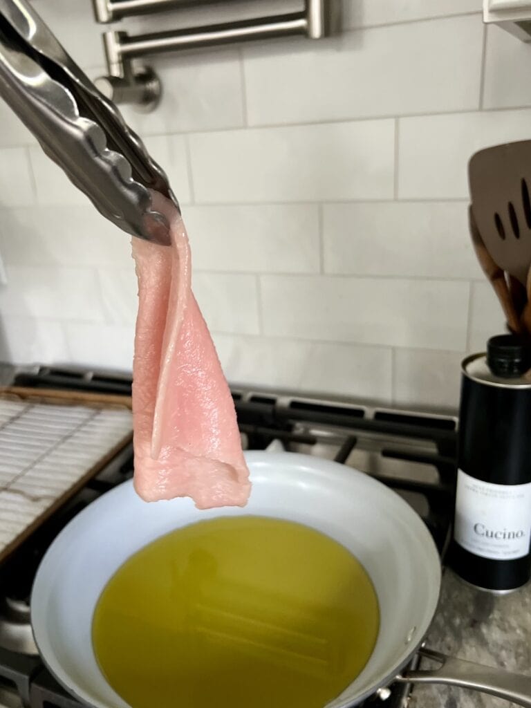 Holding up a piece of thin pork loin with tongs to show its thickness, with frying pan with oil in background.