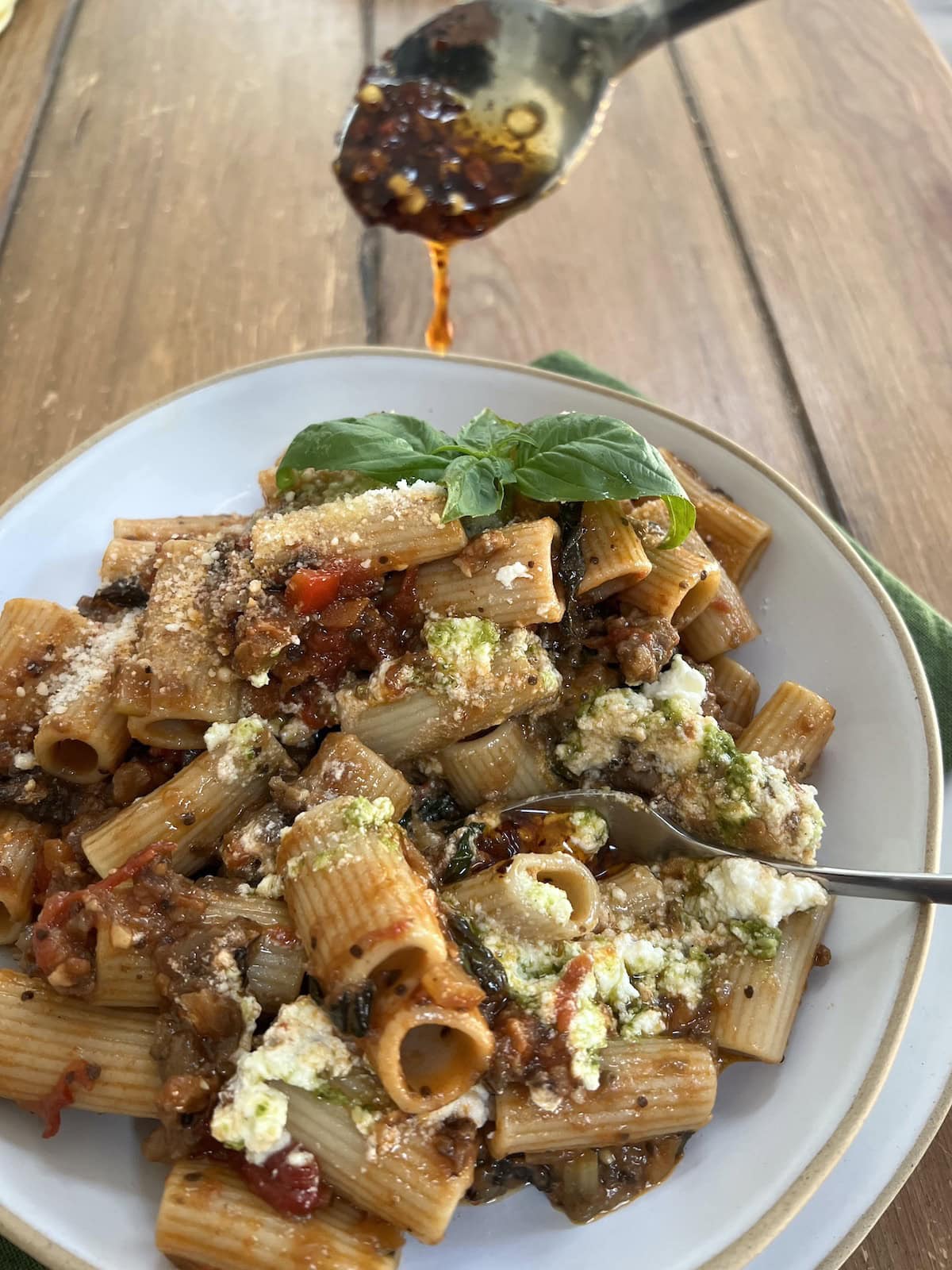 Drizzling spicy pepper oil (olio santo) into Eggplant Sausage Pasta with basil garnish in pasta bowl.