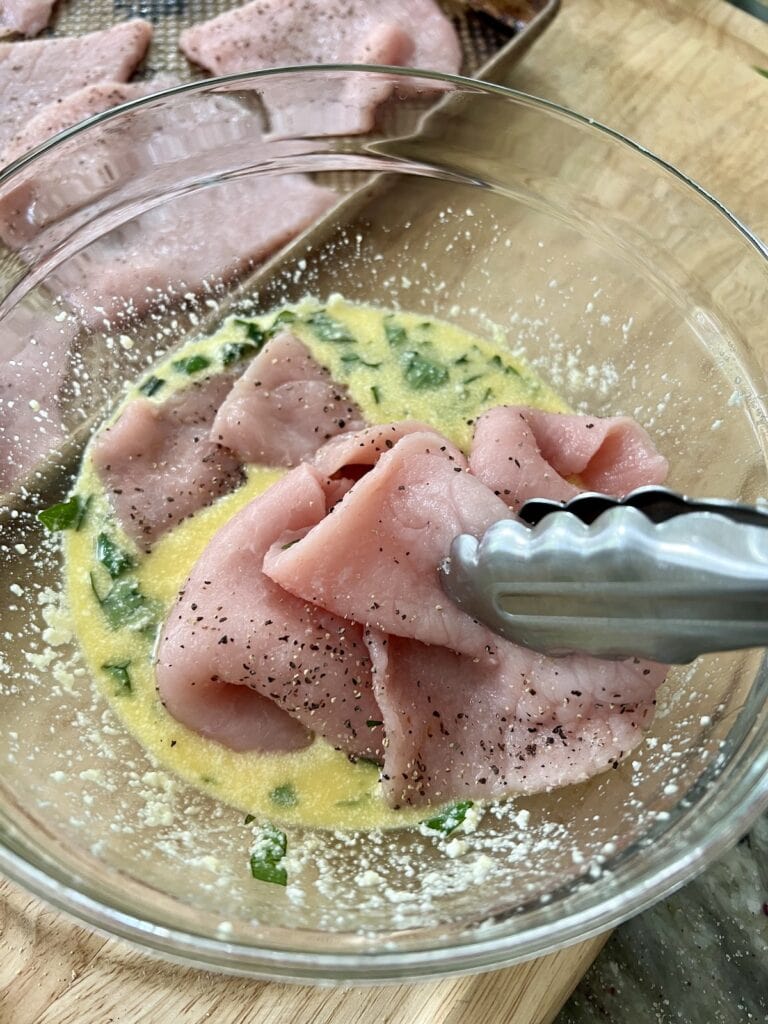 Placing thin pieces of seasoned pork into egg, milk herb and cheese mixture.