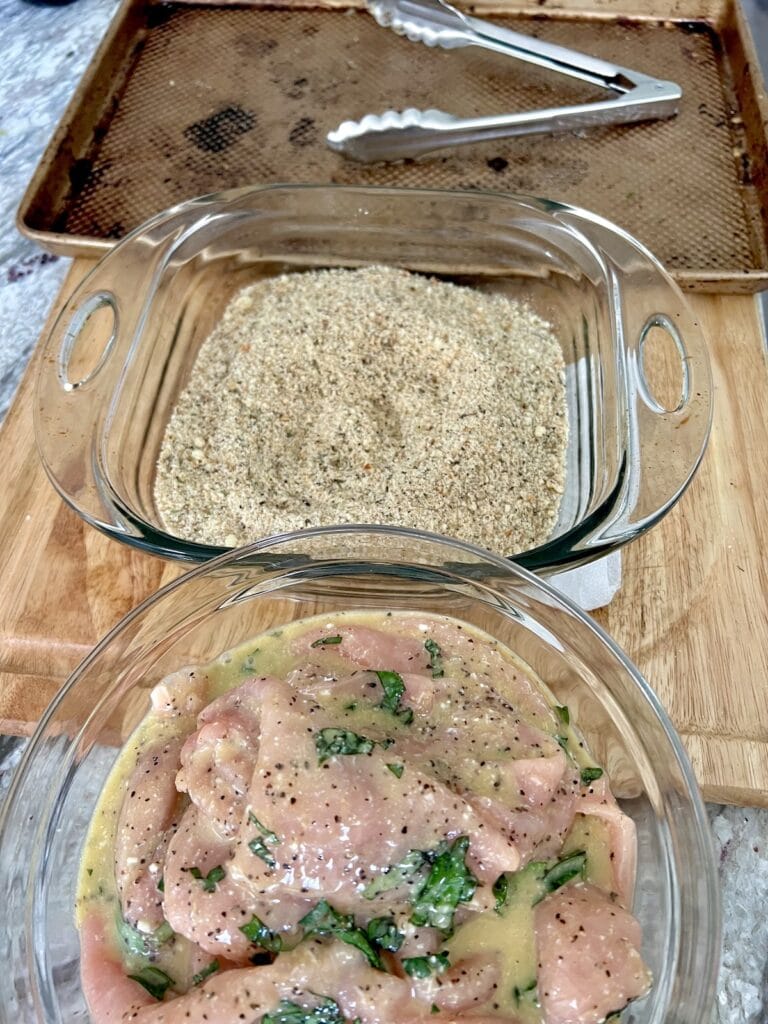 Bowl of thin pork in seasoned egg and milk mixture next to a pan of seasoned breadcrumbs next to an empty sheet pan with tongs on it.