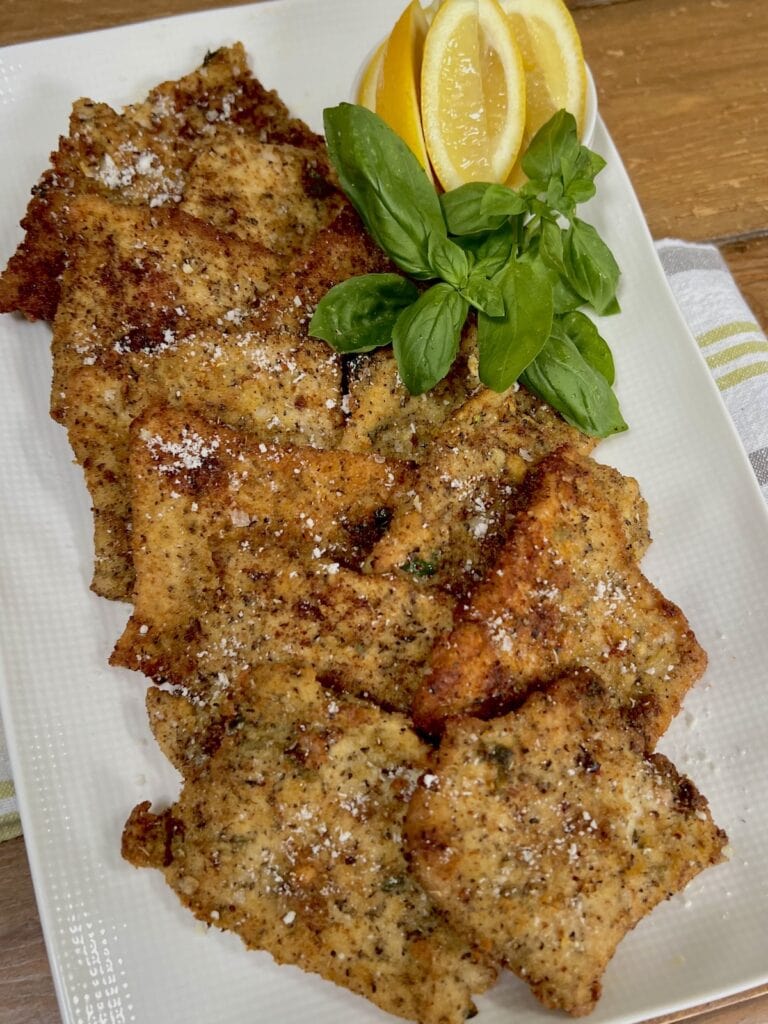 Several crispy pork cutlets on white serving platter with lemon wedges and basil garnish.